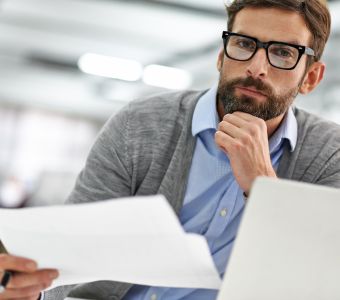 BerufsWege für Frauen und Social Business Women e.V., Weiterbildung für Frauen, WS WiEPa Risiken systematisch betrachten, ein Mann sitzt an seinem Laptop, Unterlagen in der Hand und schaut nachdenklich in die Kamera