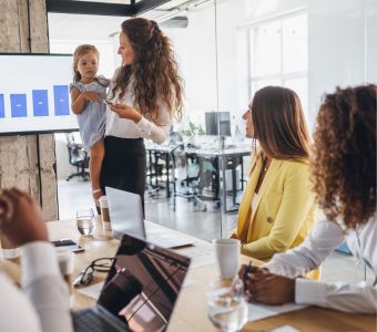 BerufsWege für Frauen und Social Business Women e.V., Weiterbildung für Frauen, WS WiEPa Sichtbarkeit und Positionierung nach der Elternzeit, Frau im Büro hat ihr Kind auf dem Arm und hält Präsentation vor Team