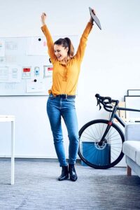 Frau in gelber Bluse mit Fahrrad jubelt und tanzt im Büro-Female-Empowerment_MKK-Berufswege-für-Frauen-e.V.