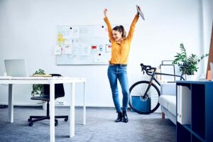 Frau in gelber Bluse mit Fahrrad jubelt und tanzt im Büro-Female-Empowerment_MKK-Berufswege-für-Frauen-e.V.