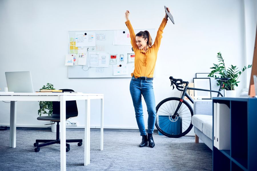 Frau in gelber Bluse mit Fahrrad jubelt und tanzt im Büro-Berufswege-für-Frauen-e.V.
