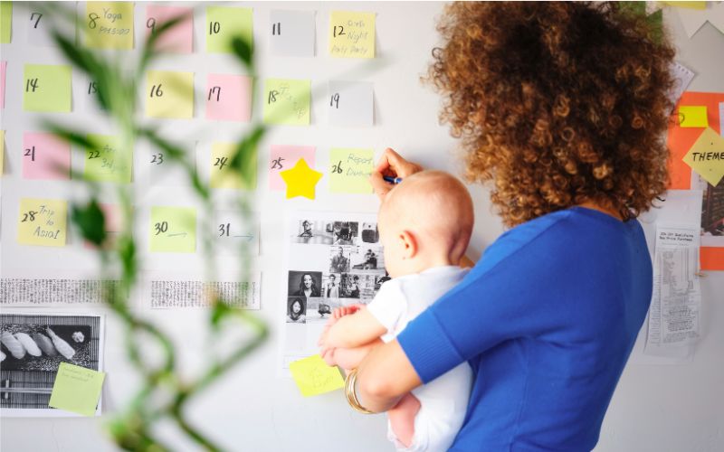 WS Ordentlicher Wiedereinstieg_Weiterbildung BerufsWege für Frauen e. V., Eine Frau mit Baby im Arm steht vor ihrer Terminplanung, es herrscht eine freundliche und positive Atmosühäre